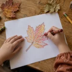 Child coloring a hand-drawn autumn leaf beside real fallen leaves on a wooden table